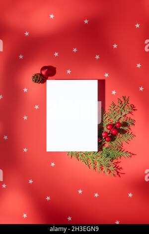 Composizione di Natale. Foglio rettangolare di carta bianca su sfondo rosso con stelle e tenui ombre, decorato con ramoscelli di pino, bacche rosse e CH Foto Stock