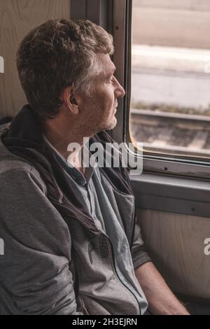 Un uomo di mezza età serio e bello è seduto nel carrello di un vecchio treno elettrico e guarda con attenzione fuori la finestra. Lavorare in un'altra città. Foto Stock