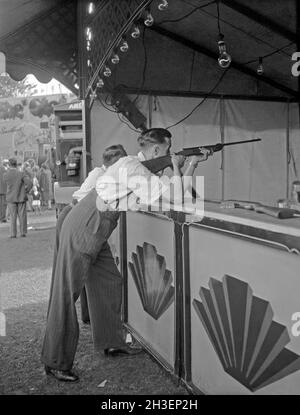 Una gamma di fucili o una galleria di tiro a un funfair nel Regno Unito c.1950 – qui un uomo prende il mira a un bersaglio con un fucile ad aria. Un buon punteggio o Bullseye guadagnerebbe un premio. Notate l'arte della fiera sotto forma di motivi decorativi 'inferno' sullo stand. Questo è tratto dal negativo in bianco e nero di un fotografo dilettante – una fotografia d’epoca degli anni ’50. Foto Stock