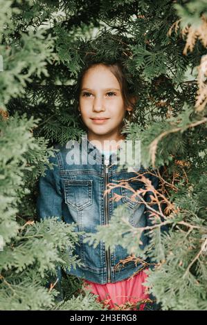 ritratto del volto di una bella caucasica caucasica candida sana bambina di otto anni circondato da rami e foglie di verde thuja pianta o cypres Foto Stock