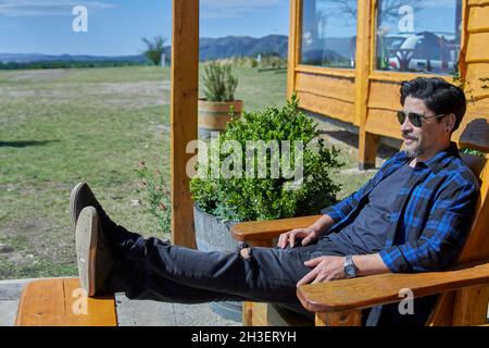 uomo latino seduto su una sedia di legno rilassante in un vigneto in attesa di una visita guidata. Córdoba, Argentina. Orizzontale Foto Stock