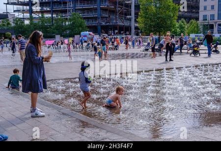 I genitori guardano i propri figli e si rilassano sulle panchine mentre i bambini giocano in acqua presso le fontane di Granary Square, King’s Cross, Londra. Foto Stock