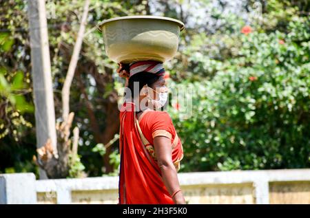 SURAJPUR, INDIA - 08 ago 2021: Una vista laterale di una donna di mezza età in una maschera che porta un contenitore di plastica sulla testa con verdure in vendita Foto Stock