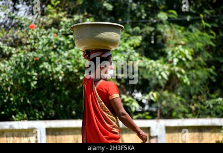 SURAJPUR, INDIA - 08 ago 2021: Una vista laterale di una donna di mezza età in una maschera che porta un contenitore di plastica sulla testa con verdure in vendita Foto Stock