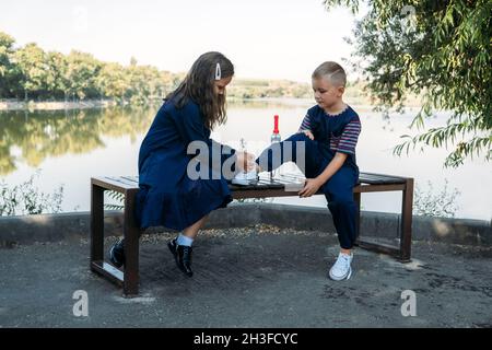 La bambina aiuta ragazzo fratello cravatta scarpe su panca in parco. Sorella cravatta merletti su scarpe fratello. Ragazza amico insegna il bambino a legare le scarpe. Amore Foto Stock