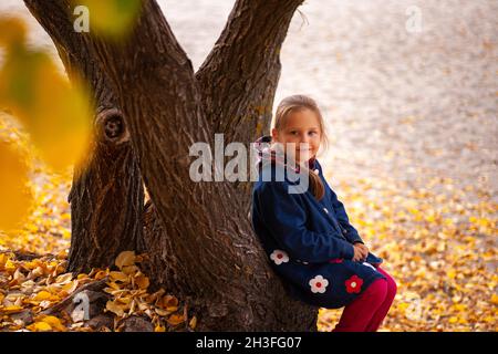 Il bambino in giacca blu è seduto su un moncone. La bambina ama passeggiare attraverso la foresta autunnale, sedendosi vicino alla riva del fiume e ammirando le foglie d'autunno gialle Foto Stock