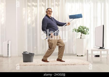 Scatto completo di un uomo maturo che balla con un mop di pulizia davanti alla tv e guarda la fotocamera Foto Stock