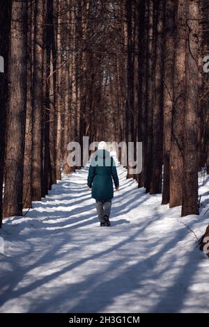 La giovane donna cammina lungo la strada. Strada invernale in foresta coperta di neve, giovane donna in verde giù giacca passeggiate in bella gelata giornata di sole Foto Stock