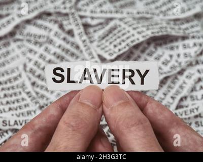 Un uomo che tiene un pezzo di carta lacerato con la parola schiavitù nelle sue mani sopra un mucchio di pezzi di carta che hanno la parola schiavitù su di loro. Chiudi Foto Stock