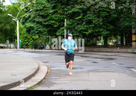 Medellin, Colombia - Dicembre 22 2020 : l'uomo latino con maschera bianca e camicia blu corre vicino allo stadio in una pista ciclabile del parco pubblico Foto Stock