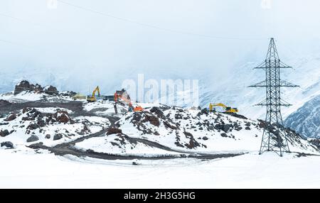 Tre escavatori stanno lavorando nelle montagne innevate accanto alla linea elettrica. Lavori di costruzione, attività in condizioni difficili. Preparazione di un sito Foto Stock