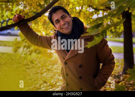 Ritratto di una discesa ispanica arabo indiano pulito rasato uomo guardando felice e sorridente mentre si guarda la macchina fotografica indossando un cappotto con sciarpa circondato wi Foto Stock
