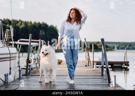 una donna felice con un grande cane bianco cammina sul molo vicino allo yacht e al mare. Foto Stock