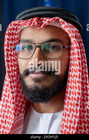 Contenuto uomo islamico con barba che indossa occhiali, agal e ghutrah guardando la macchina fotografica contro tende blu scuro Foto Stock