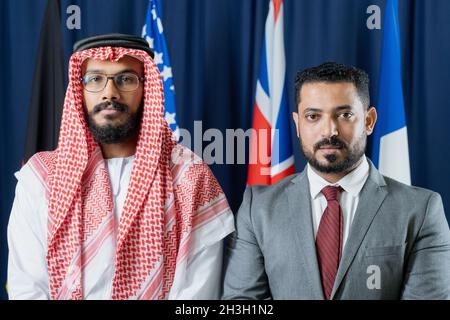 Ritratto di seri rappresentanti del Medio Oriente con barba in piedi contro le bandiere nazionali nella sala congressi Foto Stock