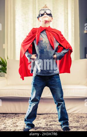 Jaunty orgogliosa giovane Super Hero in posa in un mantello rosso con le mani sui fianchi e testa in alto nel soggiorno di casa Foto Stock