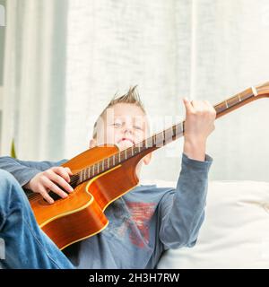 Musical giovane ragazzo strumming su una chitarra a casa come si rilassa su un divano in un primo piano alto chiave angolo basso Foto Stock