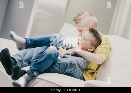 Due ragazzi ridenti che giocano su un divano a casa coccolandosi mentre si solleticano a vicenda godendo di trascorrere insieme del tempo di qualità in famiglia Foto Stock