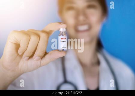 Vaccino MMR o morbillo, parotite e rosolia. Medico donna che indossa un cappotto bianco con uno stetoscopio intorno alla spalla che tiene una fiala di vetro di M-M-R va Foto Stock