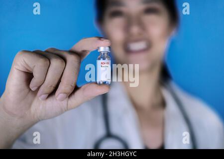 Medico donna che indossa un cappotto bianco con uno stetoscopio intorno alla spalla, tenendo un flaconcino di vetro di vaccino coniugato Pneumococcal o PCV, sorridente. Blu Foto Stock