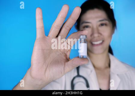 Un medico attraente, che indossa un cappotto bianco con uno stetoscopio intorno alla spalla, che tiene un flaconcino di vetro di flacone di vaccino con etichetta bianca bianca vuota, sili Foto Stock