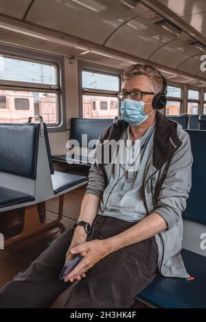 Ritratto di un uomo adulto serio in occhiali e cuffie con una maschera protettiva medica sul viso in un vagone ferroviario vuoto. Misura preventiva. Foto Stock