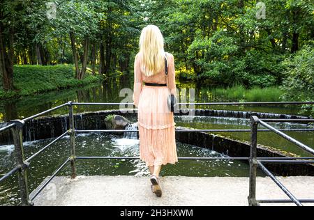 Una bella bionda alta in un abito rosa si erge sul ponte e guarda alla cascata Foto Stock