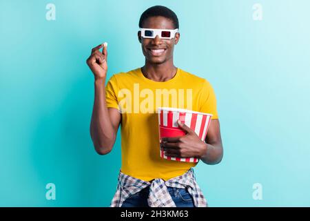 Foto di gioioso brunette hairdo giovane uomo mangiare pop corn indossare occhiali giallo t-shirt isolato su sfondo di colore ciano Foto Stock