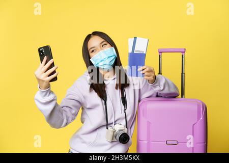 Felice donna coreana che prende selfie con passaporto e biglietti di volo, in vacanza, in piedi vicino alla grande carina valigia rosa, sfondo giallo Foto Stock