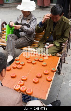 Due uomini, uno in una divisa militare, giocano / giocano a scacchi cinese gioco da tavolo in Cina mentre una donna donna donna fa il suo lavoro a maglia / maglia a Songpan, una città nella provincia settentrionale del Sichuan, Cina. (125) Foto Stock