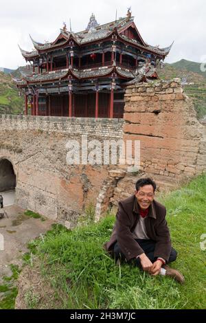 Uomo locale rilassante e seduto sull'erba fuori delle antiche mura della città / mura della città che sono stati ampiamente restaurati in grandi parti, ma con alcune sezioni non restaurate rimanendo vicino alla porta Sud visto da fuori della città di Songpan nel nord Sichuan, Cina. La casa di controllo si trova sopra l'entrata. Il locale si siede sull'erba rilassante. (125) Foto Stock