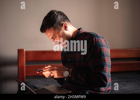 Uomo felice che usa uno smartphone e un computer portatile seduto a casa Foto Stock