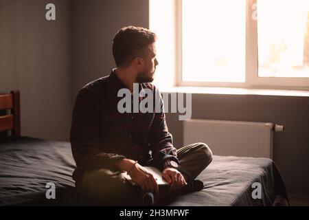 Uomo premuroso con un computer portatile che guarda attraverso la finestra seduta sul letto di casa Foto Stock