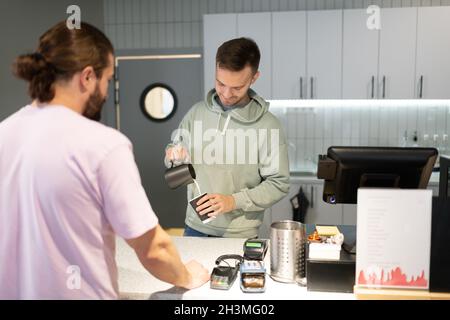 Uomo in felpa con cappuccio versando il latte dalla caraffa alla tazza mentre prepara il caffè per il cliente maschio Foto Stock