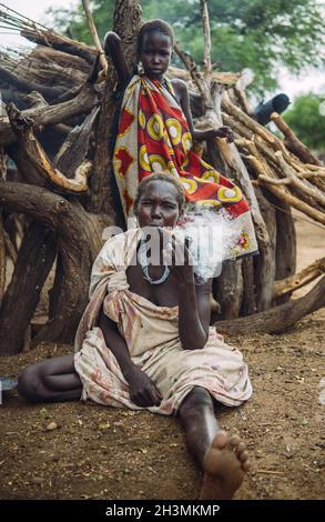 Tribù TOPOSA, SUDAN DEL SUD - 12 MARZO 2020: Donna che fuma pipa mentre si siede a terra vicino al legname e ragazza in abiti colorati Foto Stock