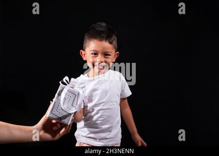Ragazzo vestito di bianco ricevendo un regalo di Natale. Isolato su sfondo nero. Felice sorridente bambino latino. Foto Stock