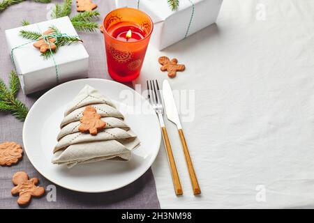 Tovagliolo in forma di albero di Natale su un piatto su tovaglia bianca con regali e decorazioni con sprigs di abete e pan di zenzero Foto Stock