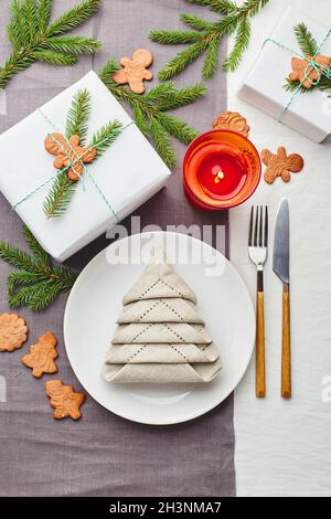 Tovagliolo in forma di albero di Natale su un piatto su tovaglia bianca con regali e decorazioni con sprigs di abete e pan di zenzero Foto Stock