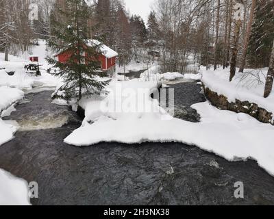 Una carina cabina di legno rosso costruita sul fiume in inverno. Coperto di neve. Foto Stock