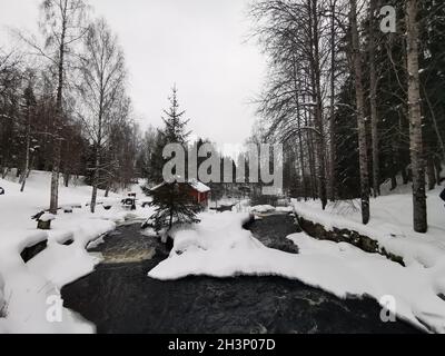 Una carina cabina di legno rosso costruita sul fiume in inverno. Coperto di neve. Foto Stock