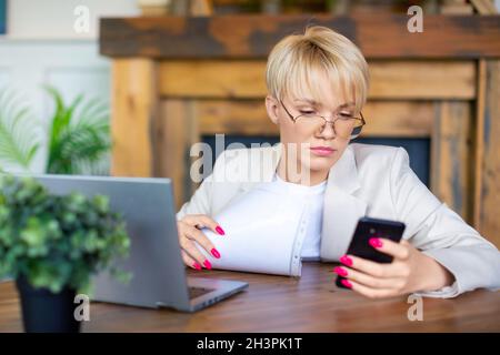 Donna bionda seria con smartphone davanti al laptop: Lavora da casa e la tecnologia moderna nel concetto di vita quotidiana. Foto Stock