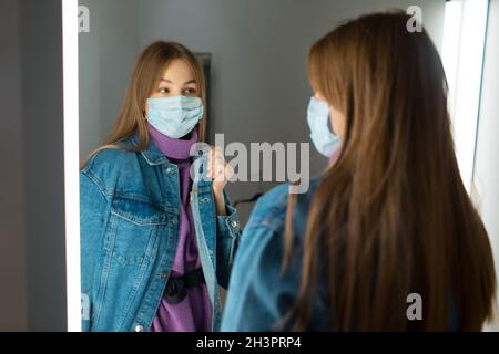 Giovane donna in maschera di faccia prova su vestiti in spogliatoio con specchio in negozio. Foto Stock