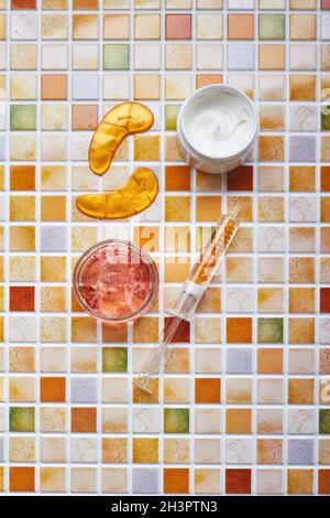 Vasetti di crema e gel per la cura del viso e del corpo, cerotti oculari in oro e siero in tubo per siringa sullo sfondo del mosaico di piastrelle. TREN Foto Stock