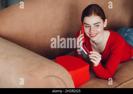 Bella giovane donna in soggiorno a casa tenendo carta di credito in mano e smartphone con box, shopping online e marketing e. Foto Stock