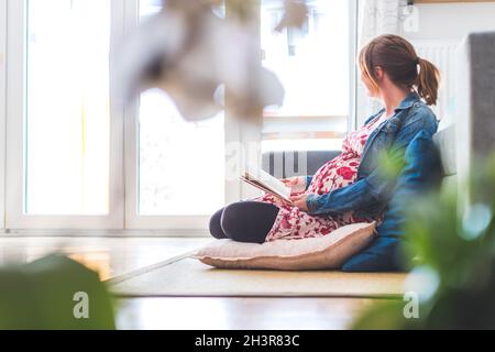 Preparazione per la gravidanza: Gravidanza la madre caucasica è seduta sul pavimento, leggendo un libro per circa i bambini Foto Stock