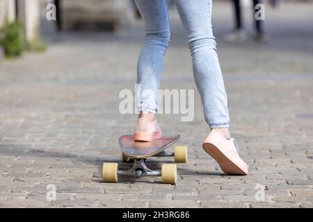 Primo piano di piedi femminili di una ragazza che indossa sneakers rosa e un jeans sullo skateboard sullo sfondo delle strade della città Foto Stock