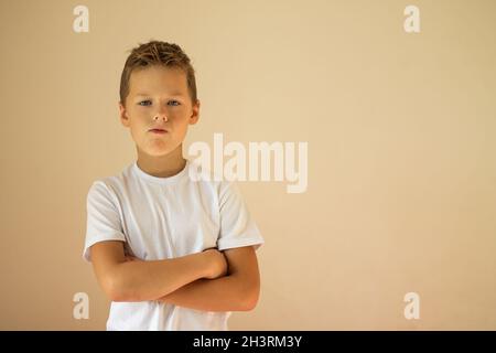 Un ragazzo serio di 7-10 anni in una T-shirt bianca si alza con le braccia incrociate su sfondo beige, spazio copia. Foto Stock
