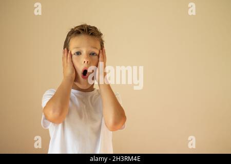 Un ragazzo sorpreso o spaventato di 7-10 anni in una T-shirt bianca si alza e grida con le mani sulle guance su uno sfondo beige. Spazio di copia. Foto Stock