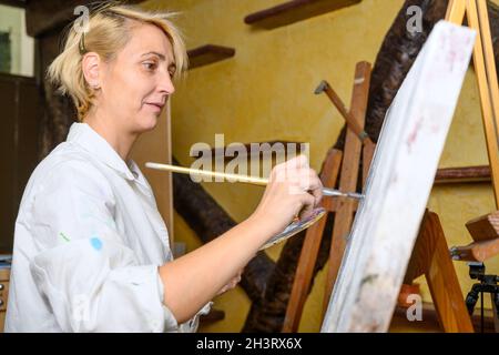 Donna felice dipinge su tela usando pennello sottile. La tela si trova sul cavalletto. L'artista disegna al cavalletto. Vista laterale della pittura femminile p Foto Stock