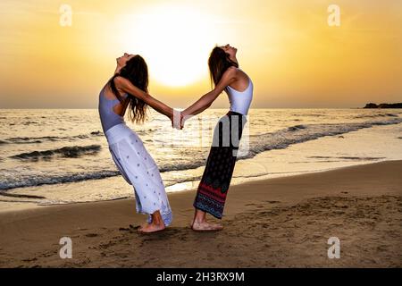 Due giovani donne di nuovo alla schiena tengono le mani e si allungano la schiena sulla spiaggia al tramonto. Senso di energia e amicizia in un'atmosfera di fine estate Foto Stock